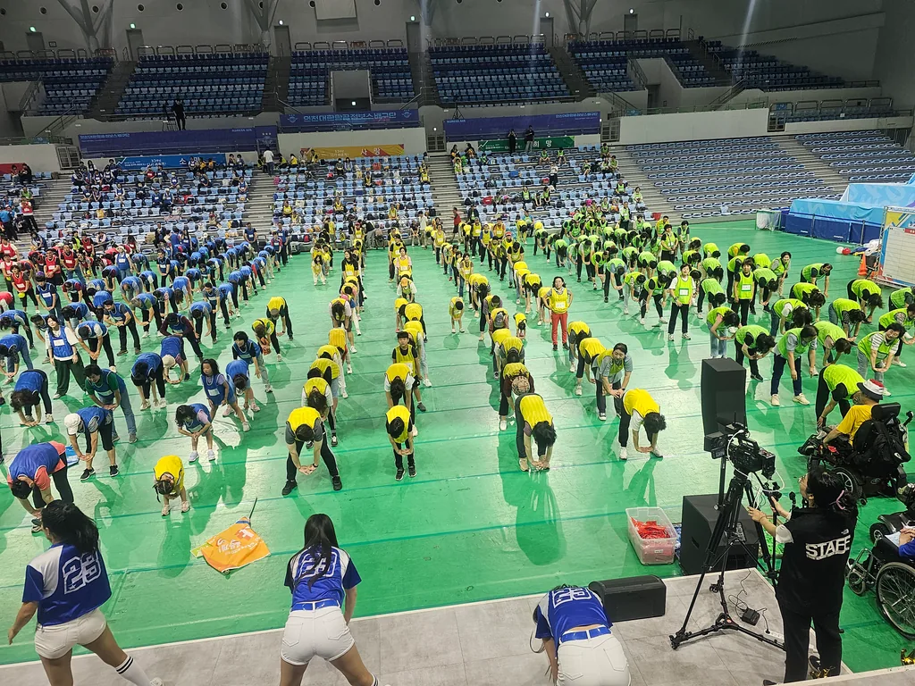 체육관 코트에서 준비운동을 이끄는 치어리더와 옆에 세운 스탠드 스피커, 촬영 카메라